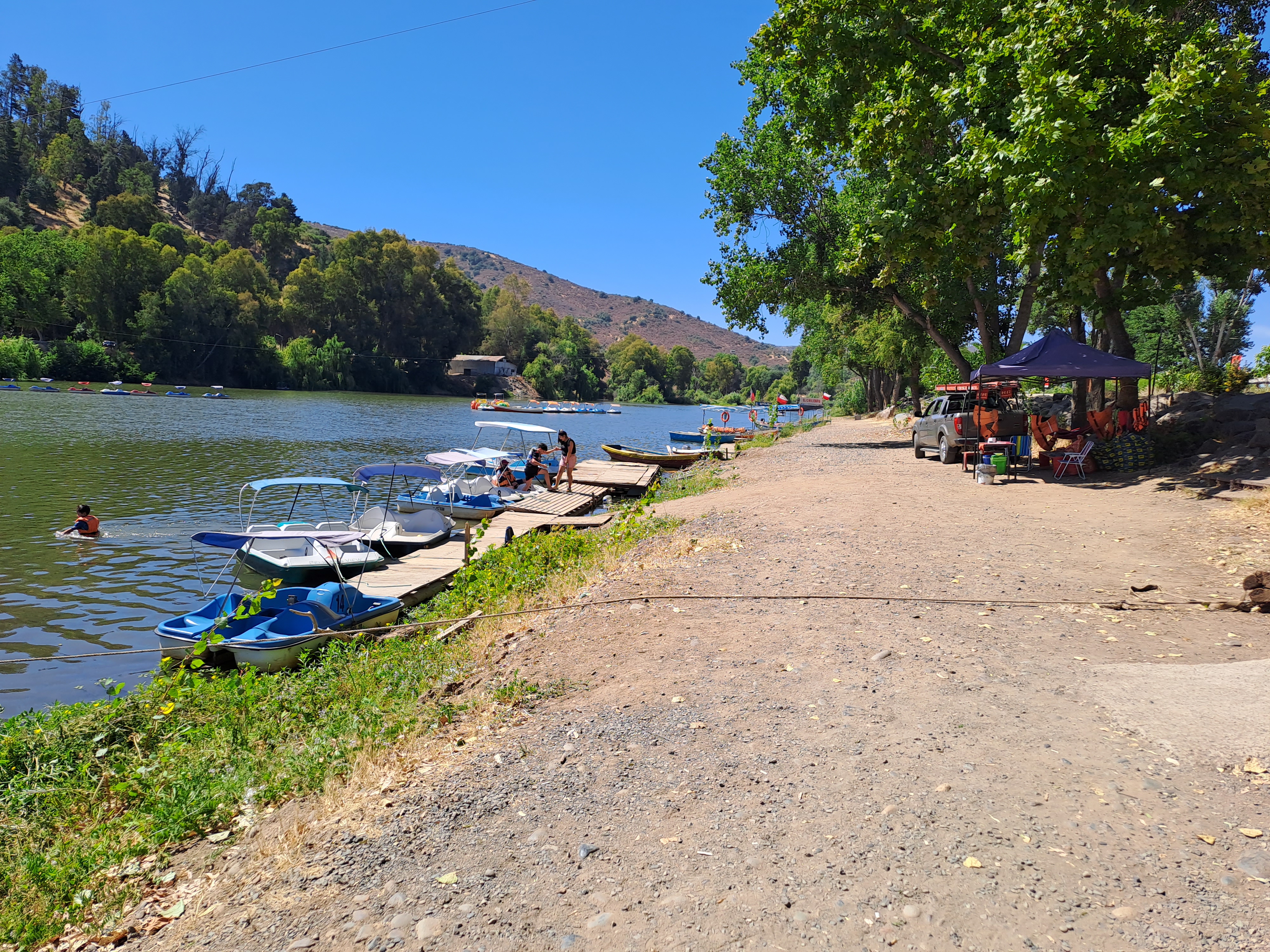 fotografía del sector del costado del río, se aprecia el sendero, aunque no es pavimentado es plano, se aprecia en el río pequeños botes para 2 personas